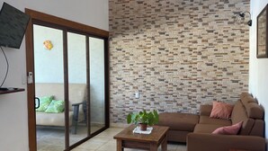 Un soggiorno con parete a effetto pietra, ventilatore a soffitto, tavolino in legno con una pianta, divano marrone e porta a vetri scorrevoli che dà su un'altra stanza.