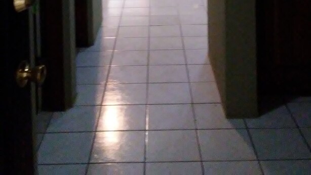 Un couloir mène à un salon équipé d’un canapé et d’un pouf, le sol est carrelé et une porte se trouve sur le côté gauche.