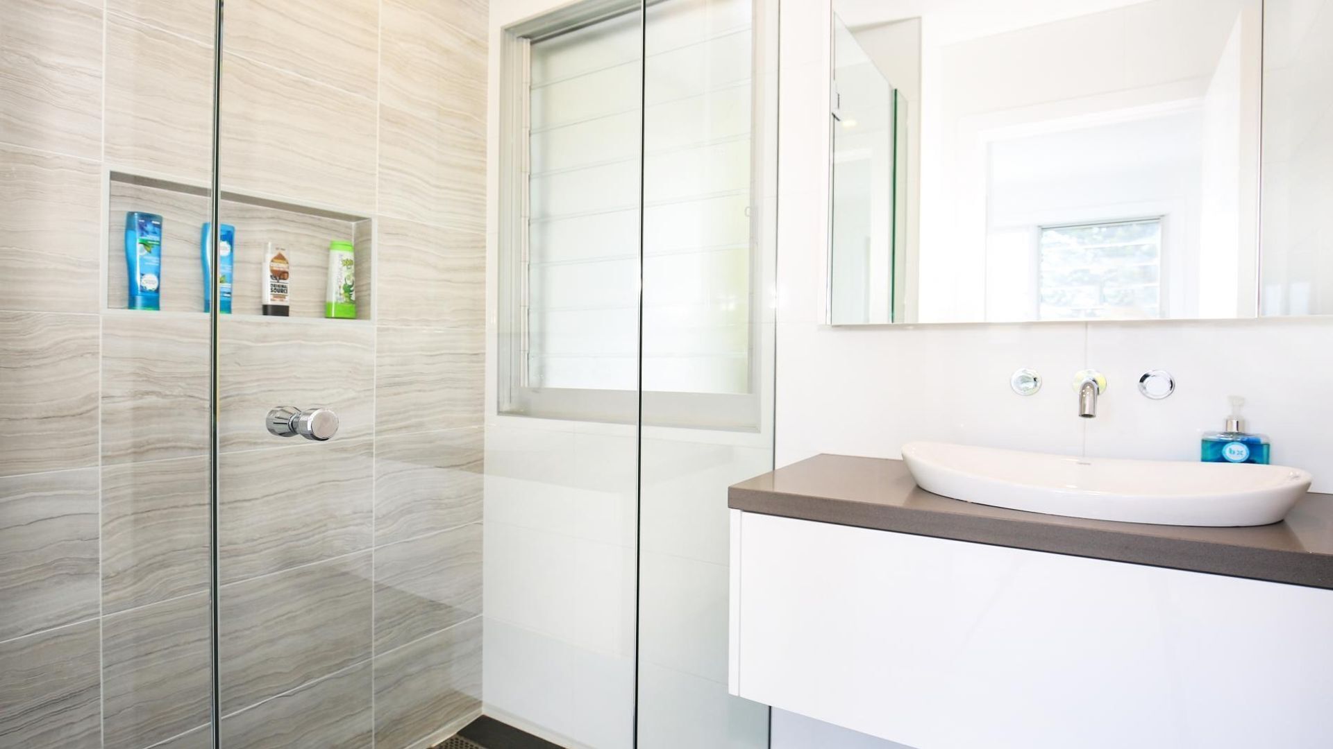 A modern bathroom with a glass shower enclosure, a white sink with a brown countertop, and a large mirror above the sink.