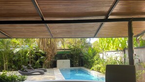 Una piscina cubierta al aire libre con una tumbona, rodeada de exuberante vegetación y un techo de madera.