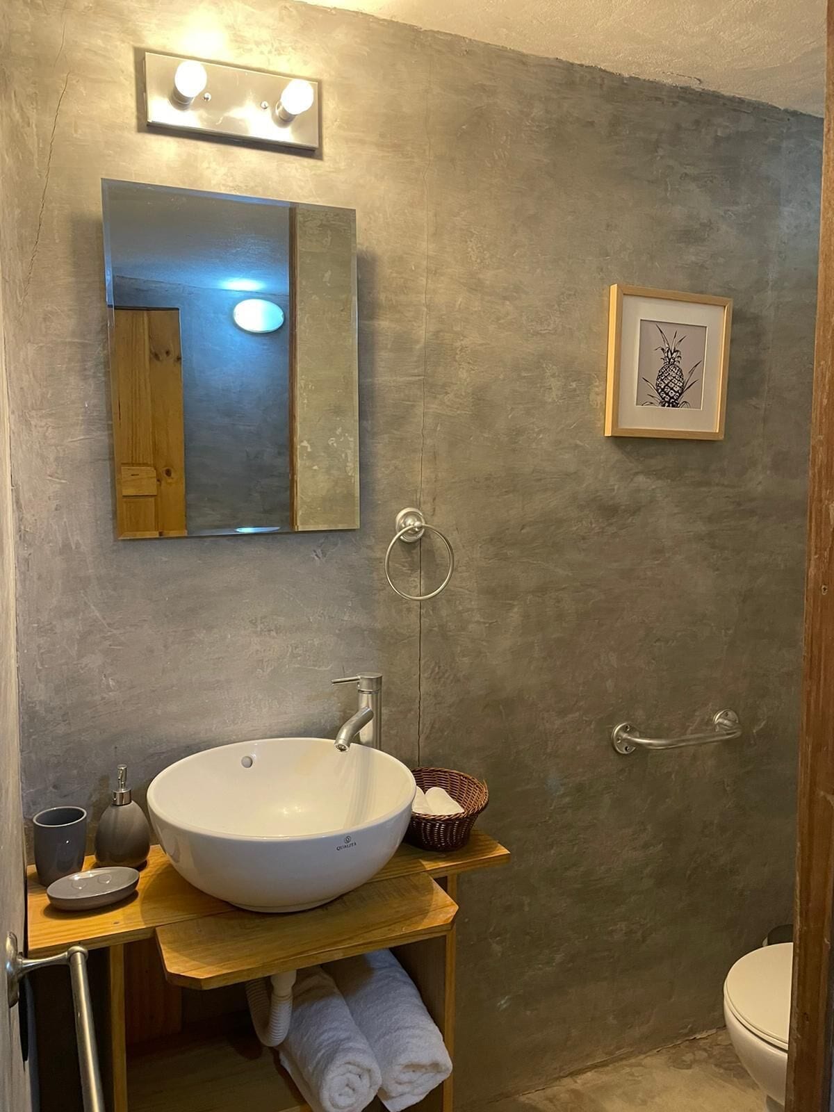 A bathroom with a large white sink, a mirror, a framed pineapple artwork, and a towel rack.
