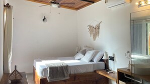 Un dormitorio con techo de madera, una cama con ropa blanca, un ventilador de techo, un equipo de aire acondicionado, una lámpara de noche y un espejo.