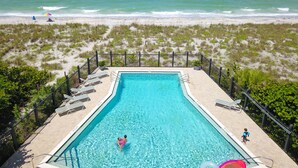 A rectangular pool with clear blue water, surrounded by a black metal fence, and a beach with sand and sparse vegetation in the background. - Tailwind South: Beautiful 3 bedroom across the street from Beach! (Placida)