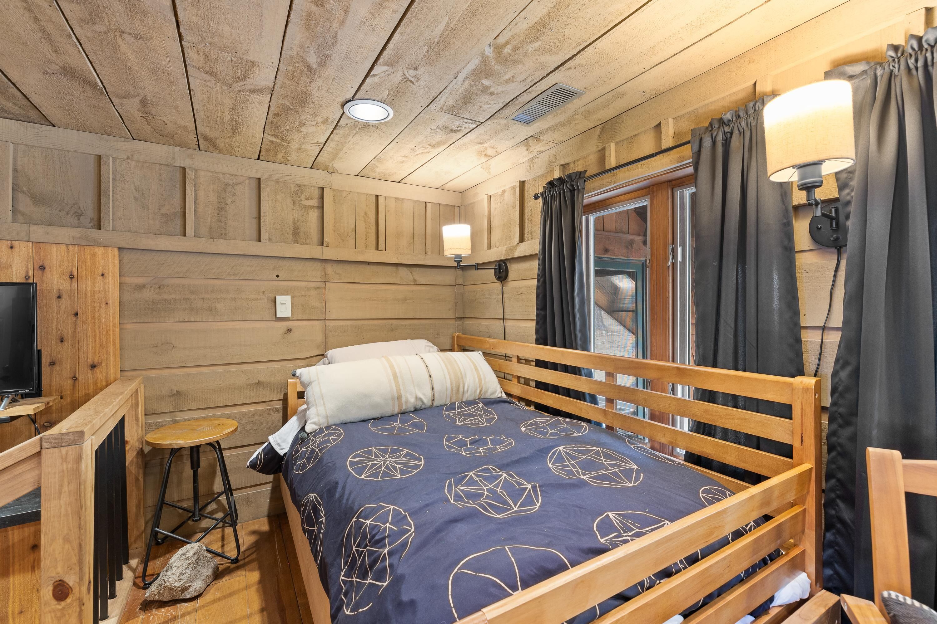 Une pièce de cabane en bois comprenant un lit, un petit tabouret, une table, une lampe et une fenêtre avec des rideaux.