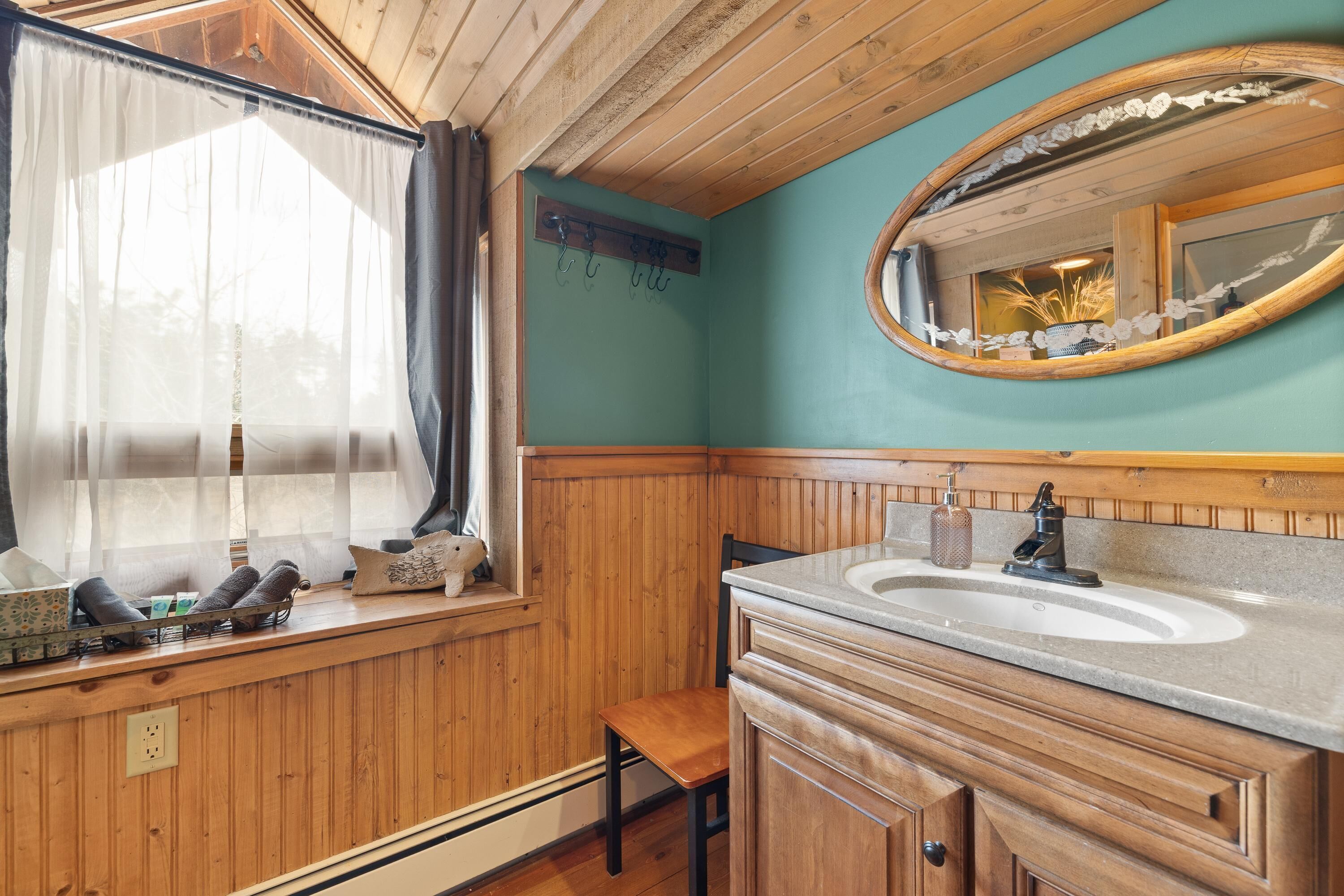 Casa de banho com painéis de madeira, uma pia, uma cadeira, uma janela com cortinas e um espelho com moldura decorada.