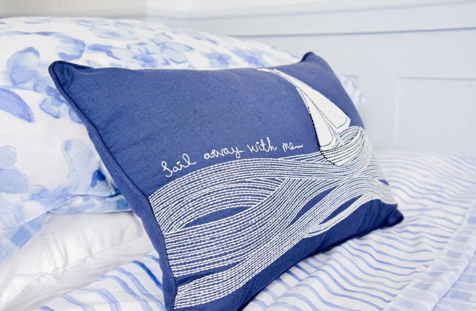 A bed with a blue pillow featuring a white wave design and the text "Sail away with me".