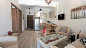 A living room with a glass coffee table, a sofa, and a TV on the wall. There is a kitchen area with cabinets and a refrigerator.