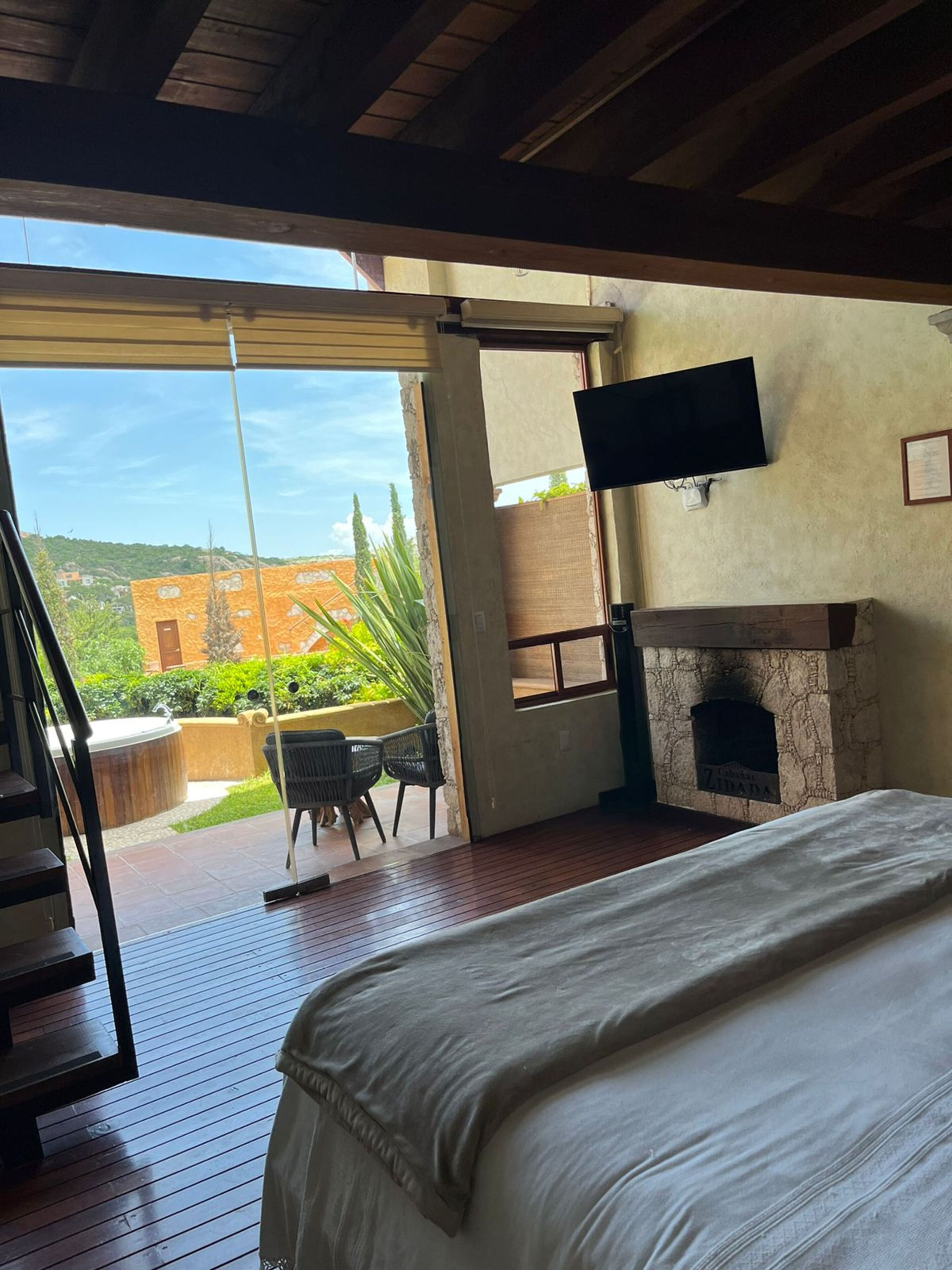A bedroom with a large bed, a fireplace, a TV mounted on the wall, and a balcony with a view of the surrounding area.