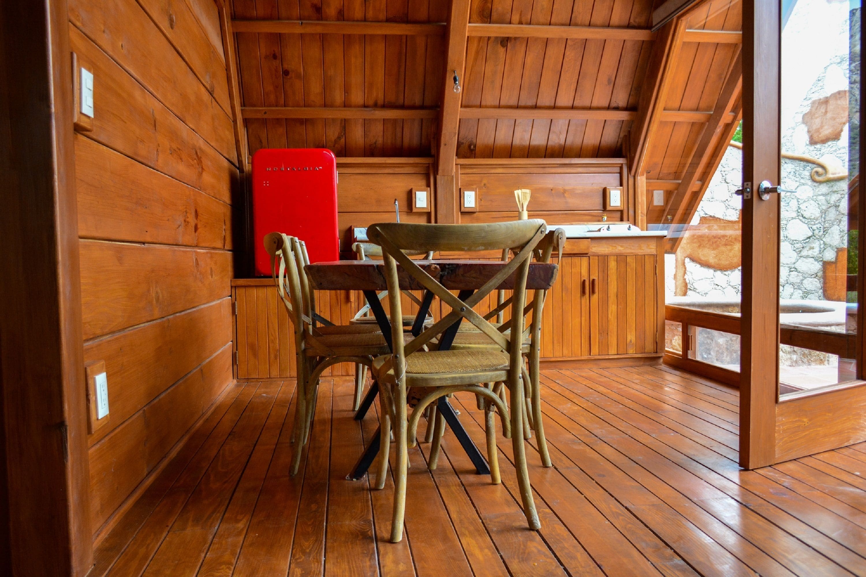 A wooden interior with a dining table and chairs, a red refrigerator, and a stone wall visible through an open door.