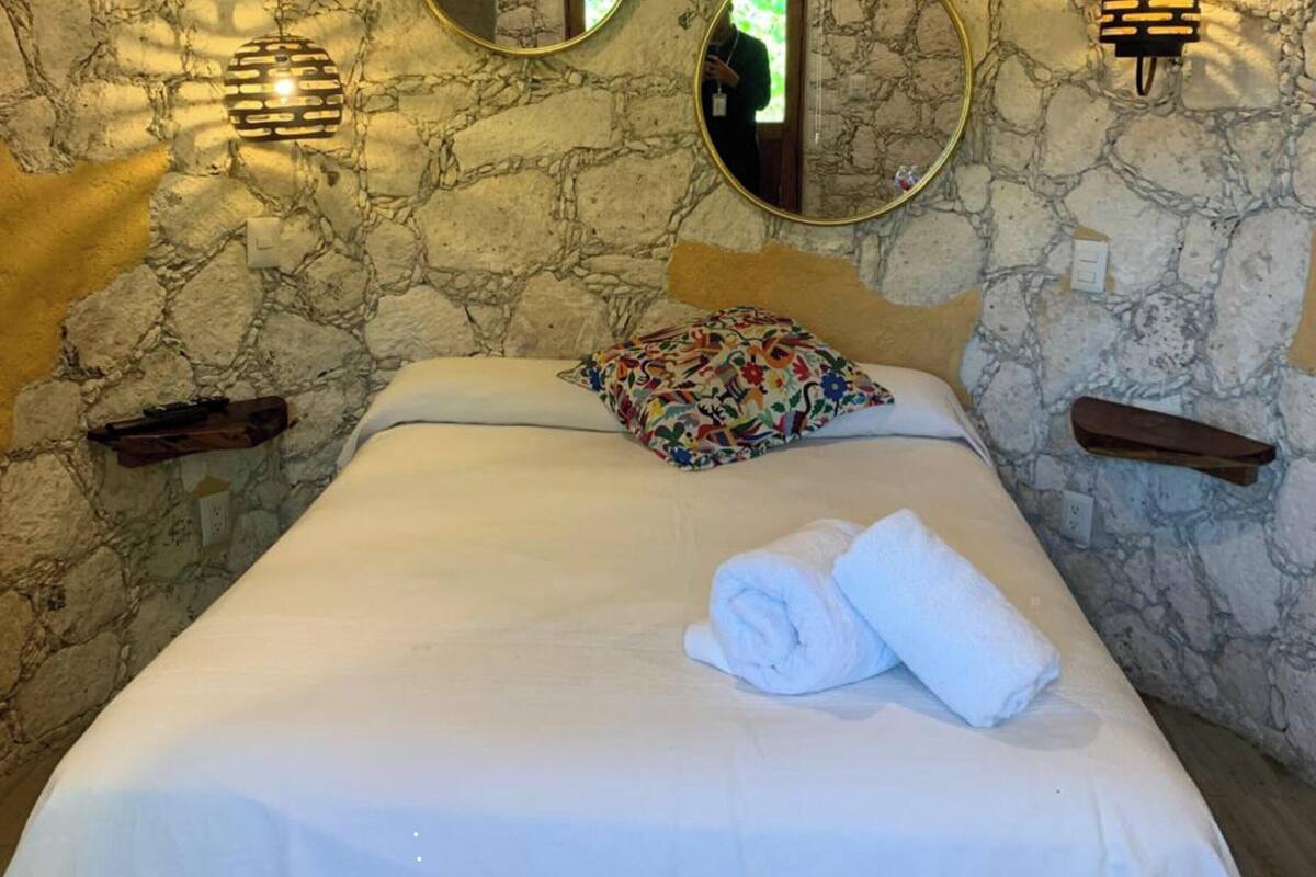 A bedroom with a stone wall, a bed with white linens, two round mirrors, and wall-mounted lights.