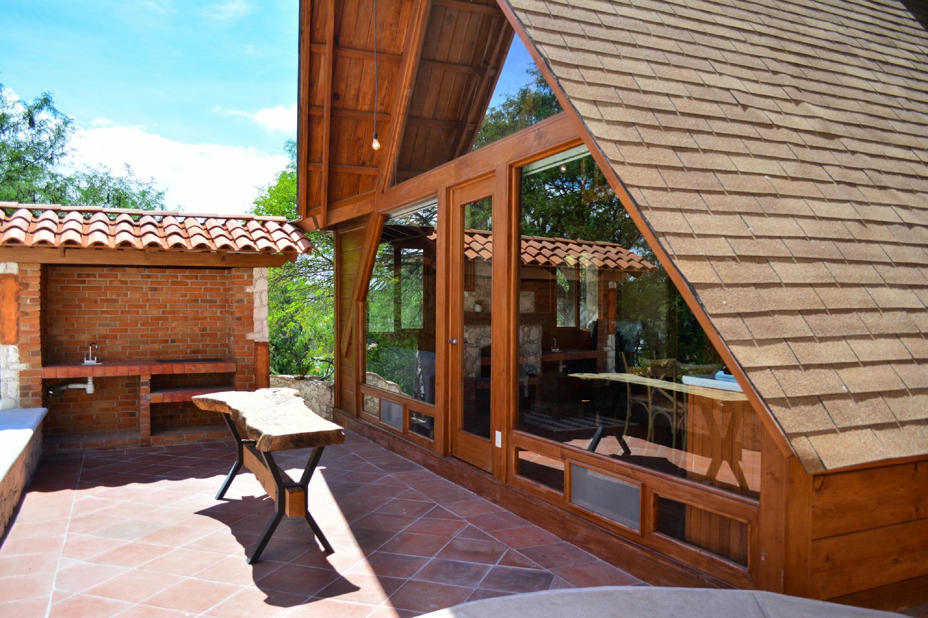 An outdoor patio with a brick barbecue, wooden tables, and large glass doors leading to a covered area.