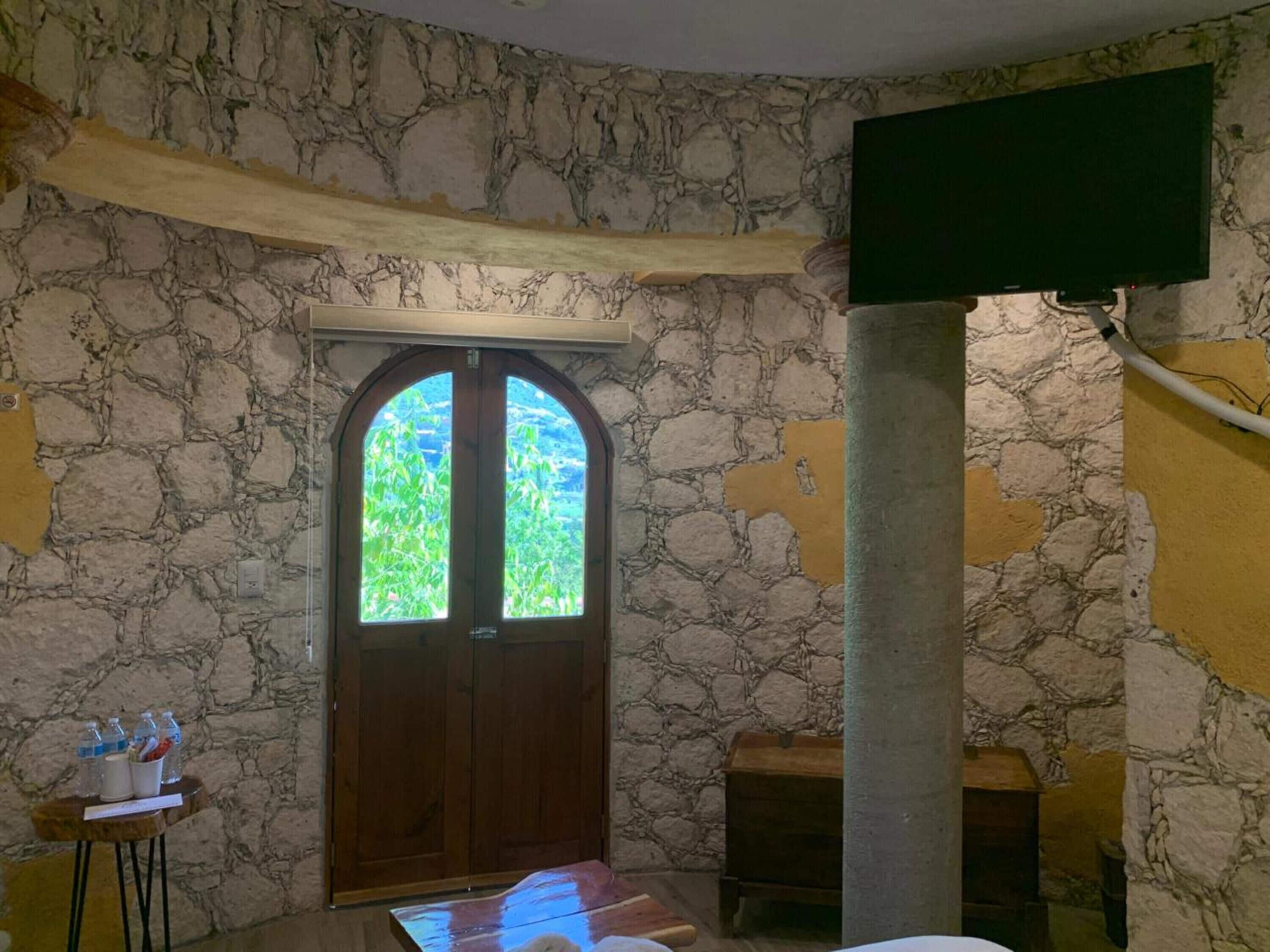 Una habitación con pared de piedra, una puerta de madera con paneles de vidrio, un televisor montado en la pared y una mesita con botellas y una taza.