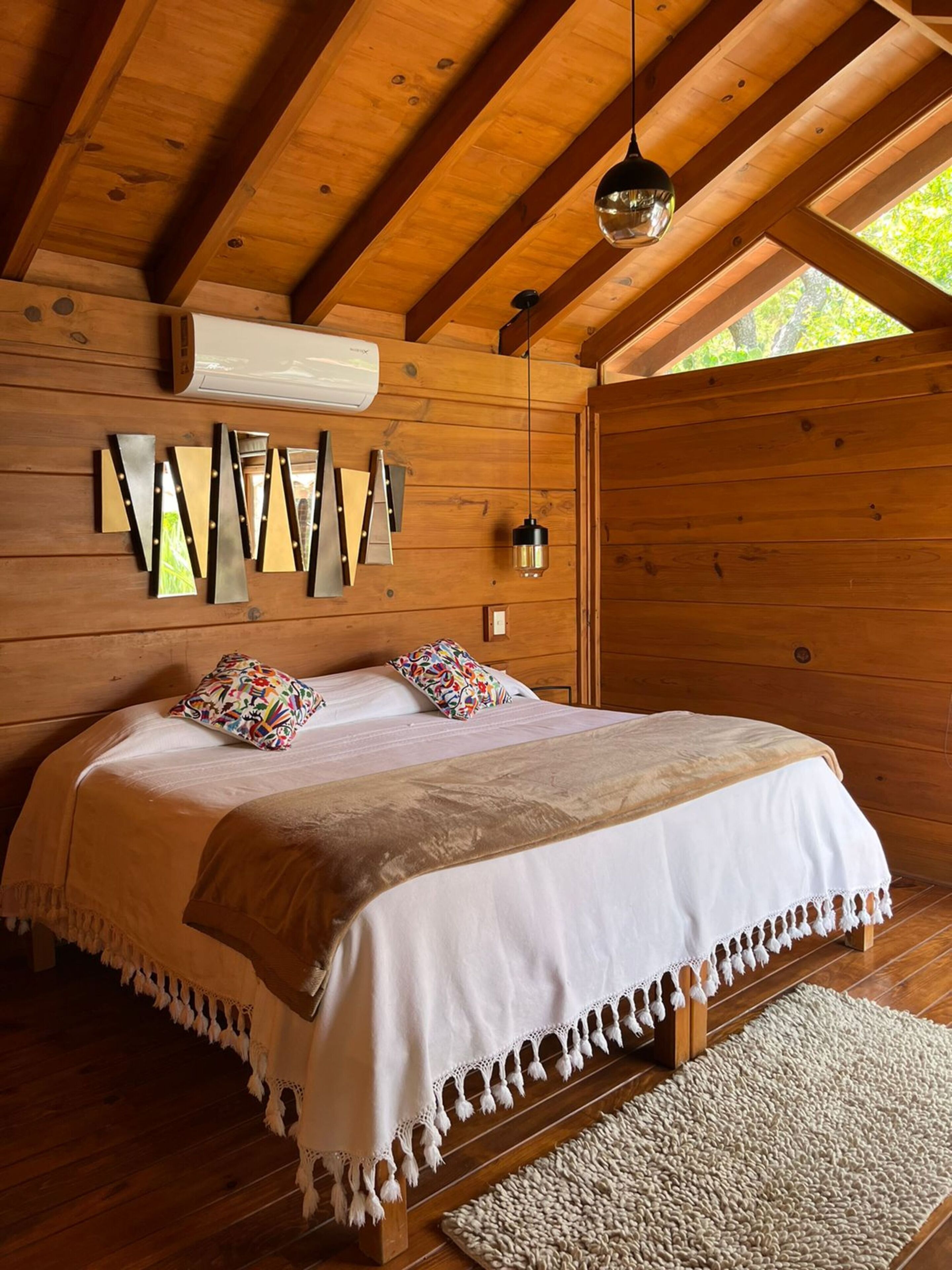 Une chambre dans une cabane en bois, avec un lit, des oreillers, un luminaire suspendu et une grille d’aération au plafond.
