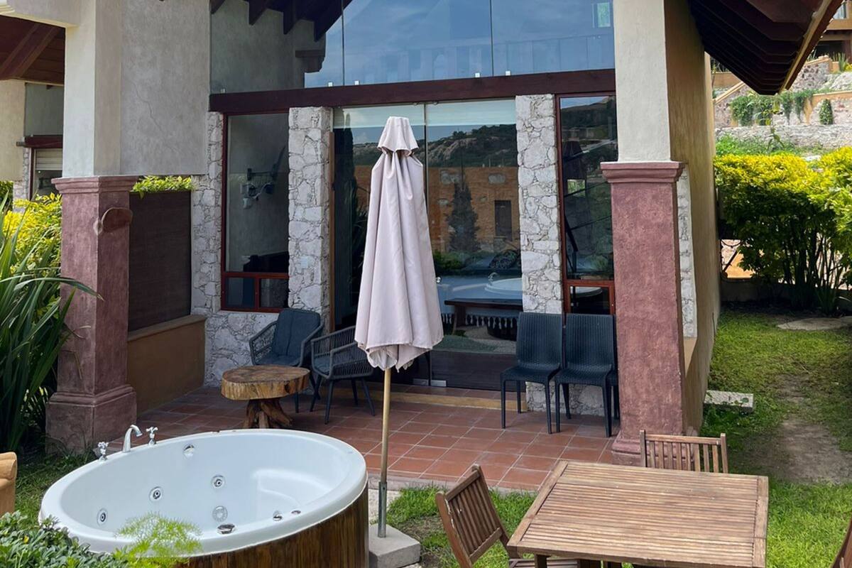 A modern house with a glass-enclosed patio, a hot tub, outdoor furniture, and a pergola.