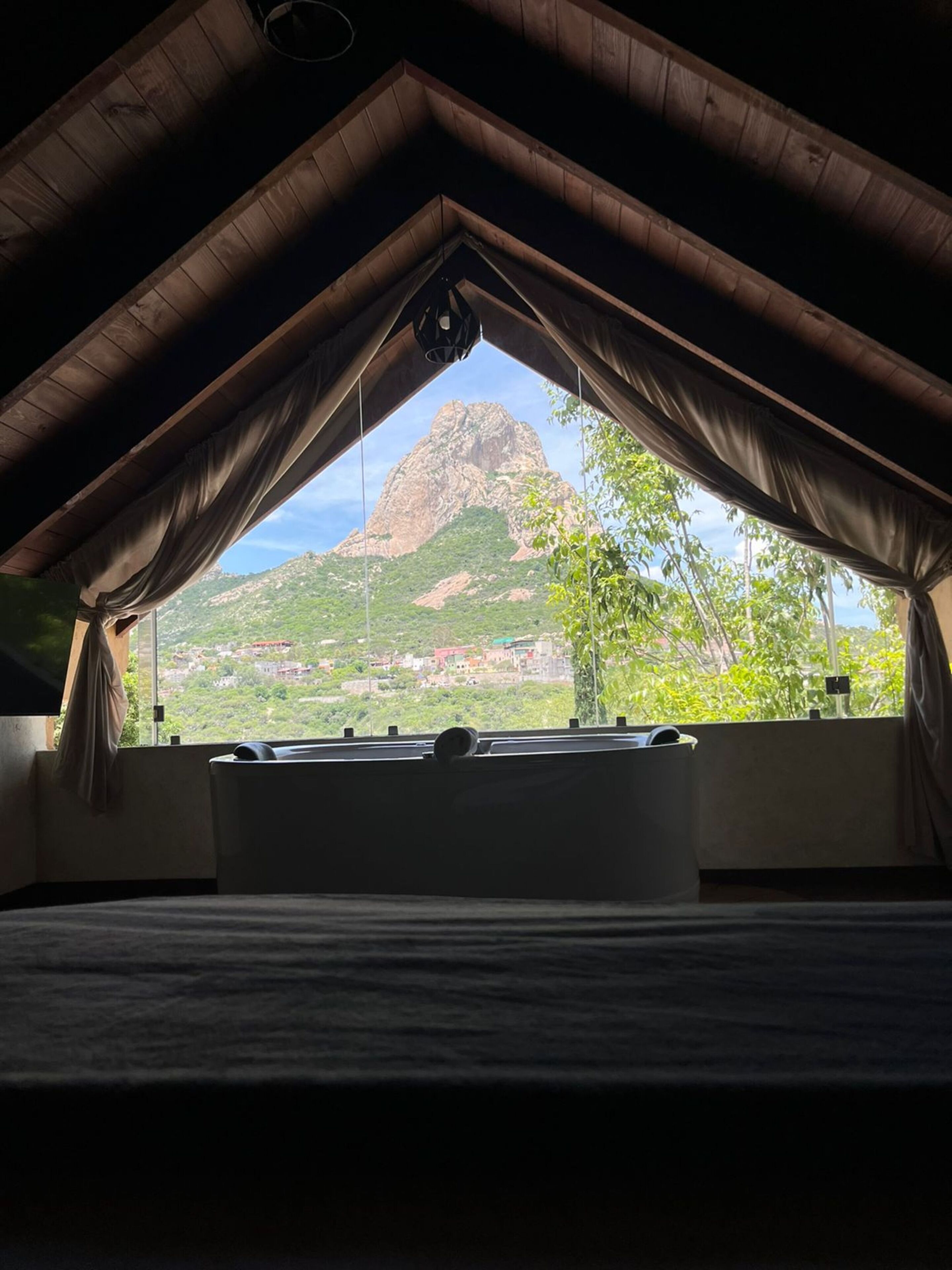 Een kamer met een schuine kap, een groot raam met gordijnen en uitzicht op een berg.