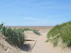 Een zandpad langs het strand dat naar de zee leidt, met schaarse begroeiing en een heldere lucht.