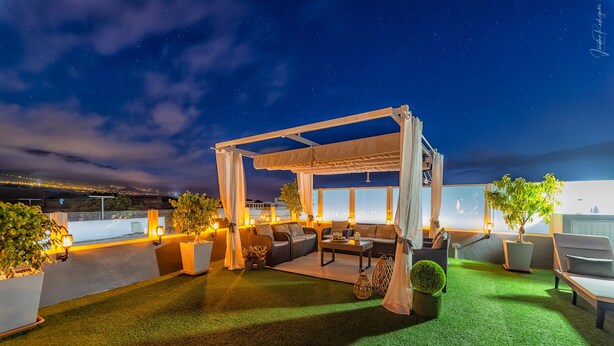 Eine Terrasse im Freien mit überdachter Sitzgruppe, Kunstrasen und Topfpflanzen.