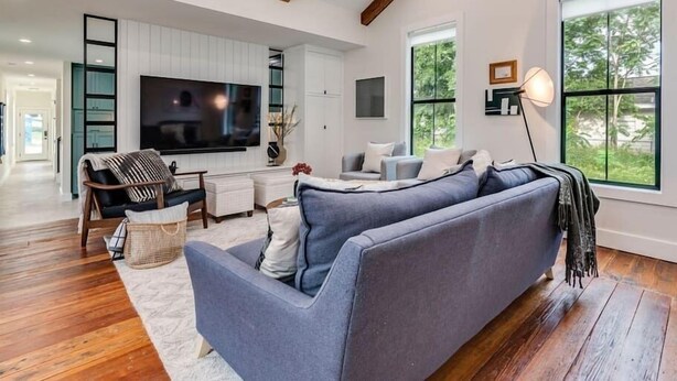 A spacious living room with a large grey sofa, wooden flooring, a flat-screen TV, and large windows providing natural light. - Modern Farmhouse with Rooftop Deck Downtown (Austin)