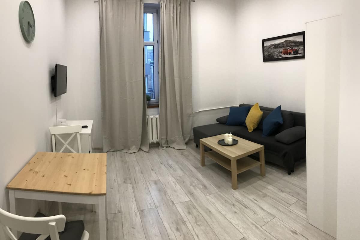 Ein kleiner, moderner Wohnraum mit einem Holztisch, einer schwarzen Couch mit Kissen, einem an der Wand montierten Fernseher und einem Fenster mit Vorhängen.