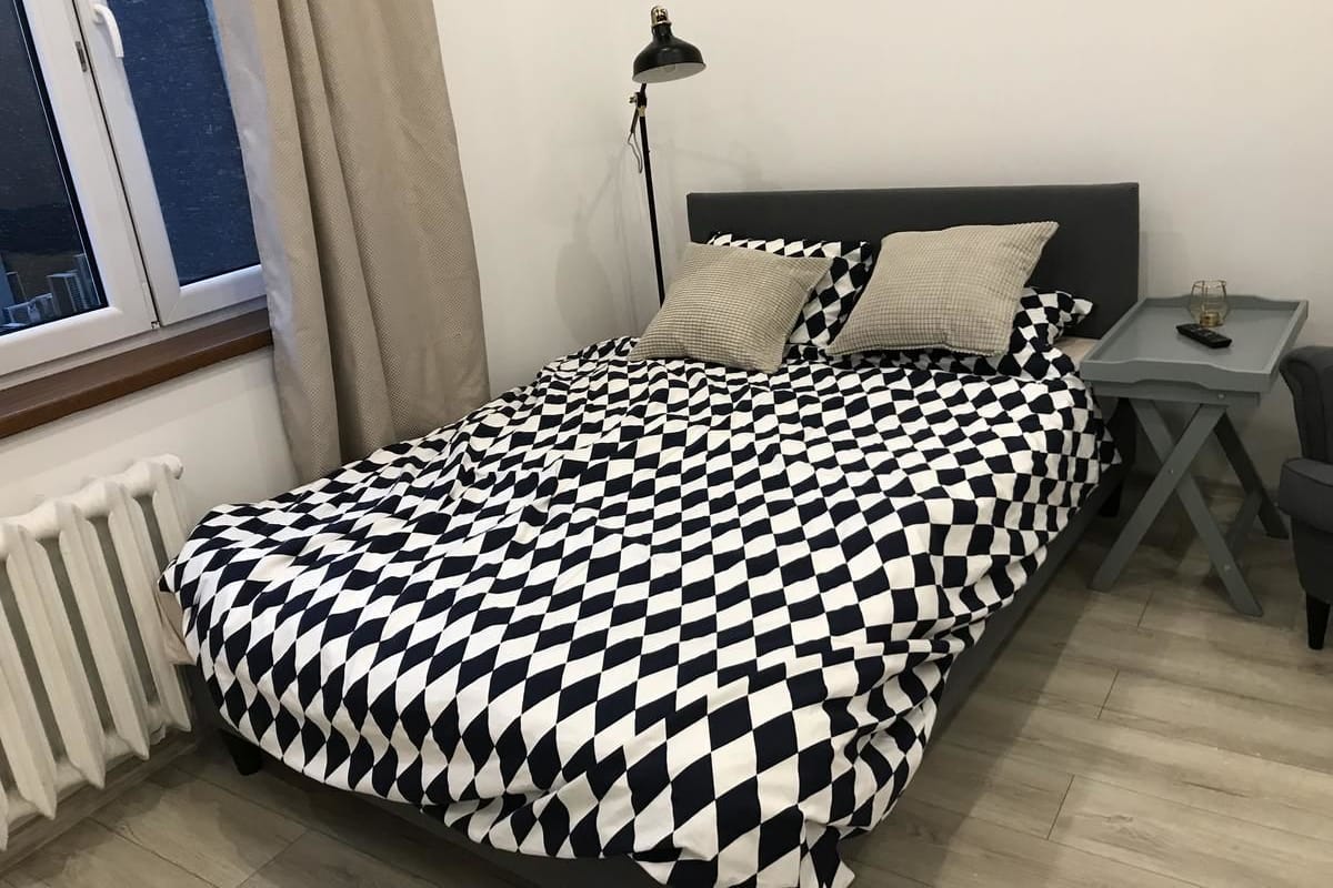 Ein Schlafzimmer mit kariertem Überwurf, einem Nachttisch mit Lampe und Blick zum Fenster.