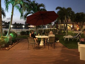 Un patio al aire libre con una mesa redonda y sillas, un paraguas grande y palmeras.