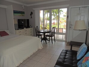 A hotel room with a bed, a TV, a dining table with chairs, a sofa, and a door leading to an outdoor area with a view of palm trees and a pool. - Aqua Mar Condos (Pompano Beach)