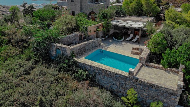 Une piscine en pierre, entourée d’une végétation luxuriante, avec vue sur la mer et un paysage montagneux en arrière-plan.