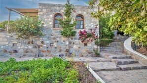Un bâtiment en pierre doté de deux fenêtres en arc, d’un escalier en pierre et d’un jardin où poussent différentes espèces de plantes.