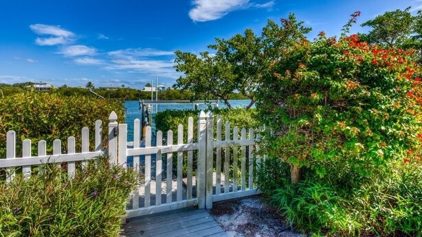 A wooden deck leading to a white picket fence, surrounded by lush greenery and a clear blue sky.