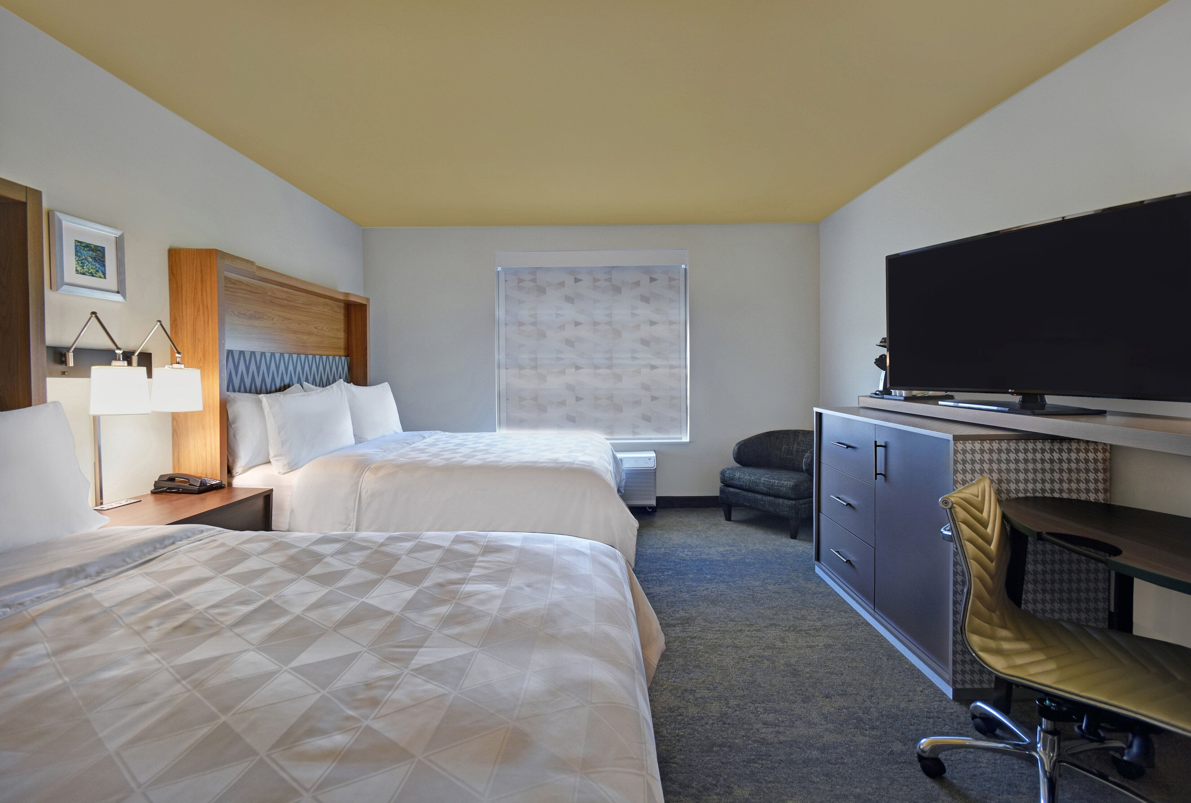 Ein Hotelzimmer mit zwei Betten, einem Schreibtisch mit Stuhl, einem Fernseher und einem Fenster mit Jalousien.