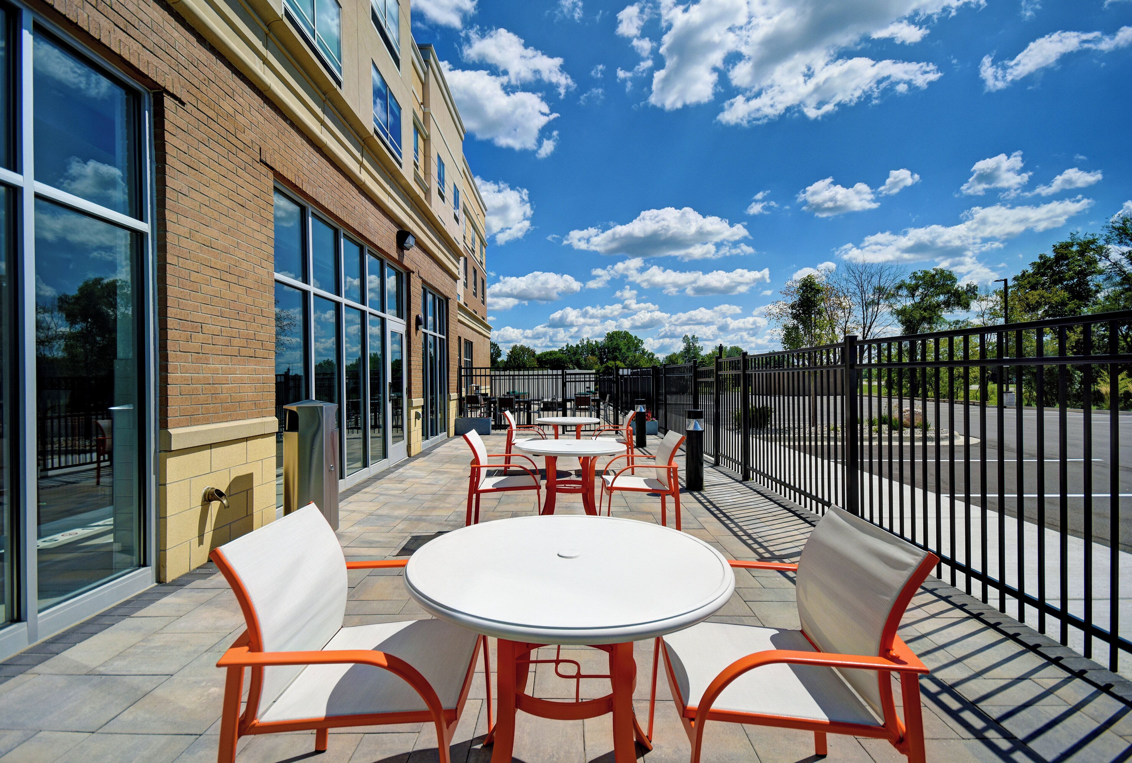 An outdoor patio with round tables and chairs, a brick building with large windows, and a clear blue sky.