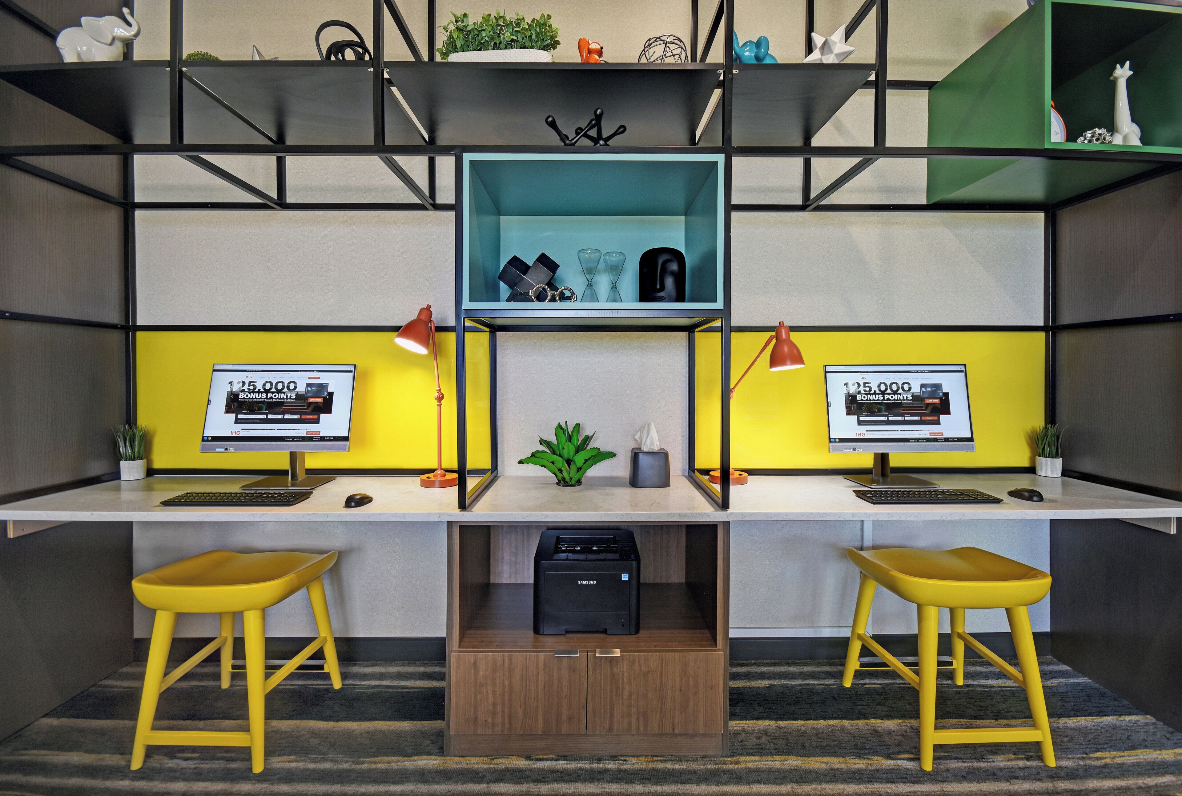 A modern office space with two desks, each with a computer monitor, keyboard, and mouse. There are two yellow stools, a printer, and decorative items on shelves.