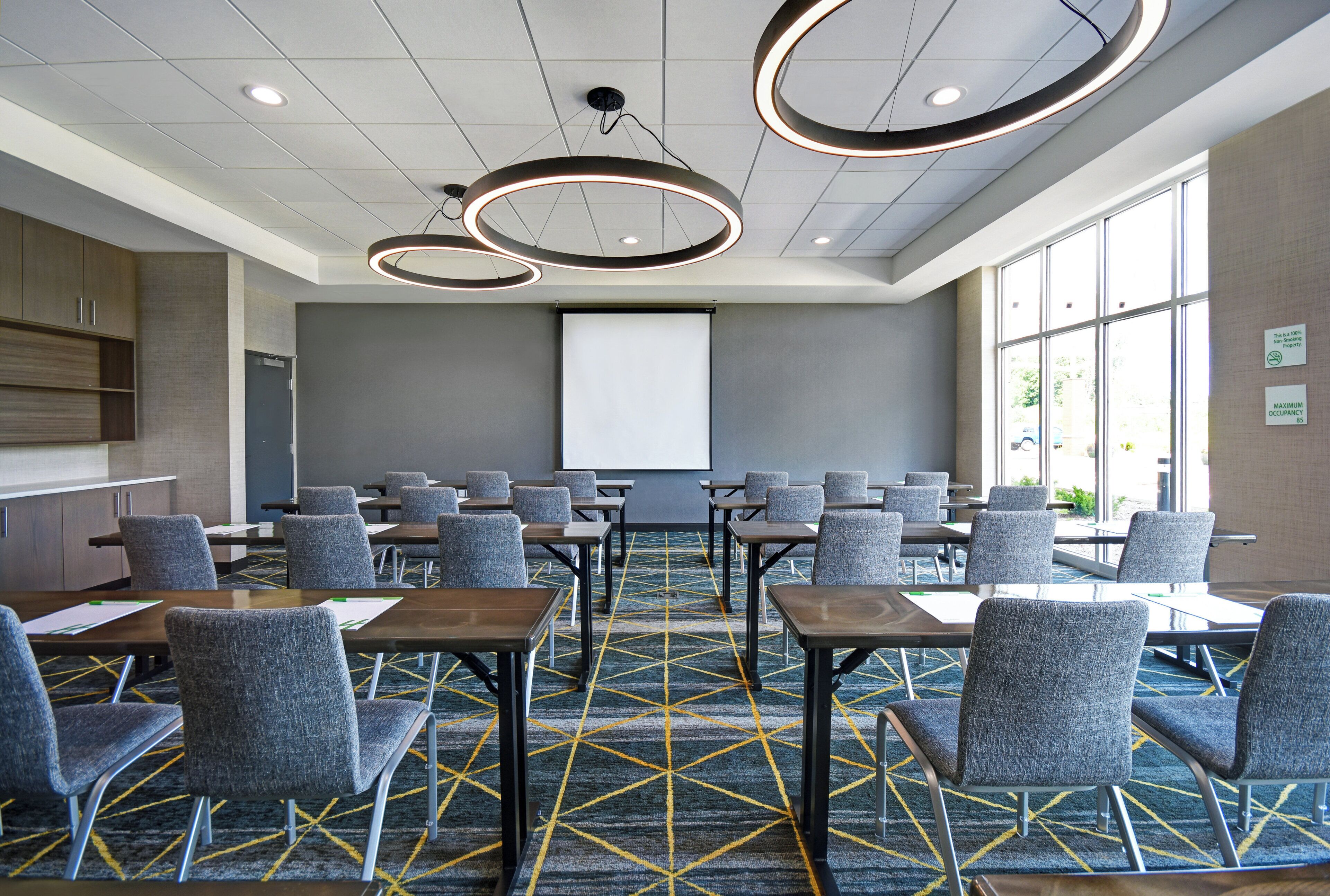 A conference room with a projector screen, rows of chairs, tables, and a patterned carpet.