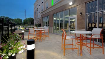 An outdoor patio area with tables and chairs, large windows, and a brick building facade.