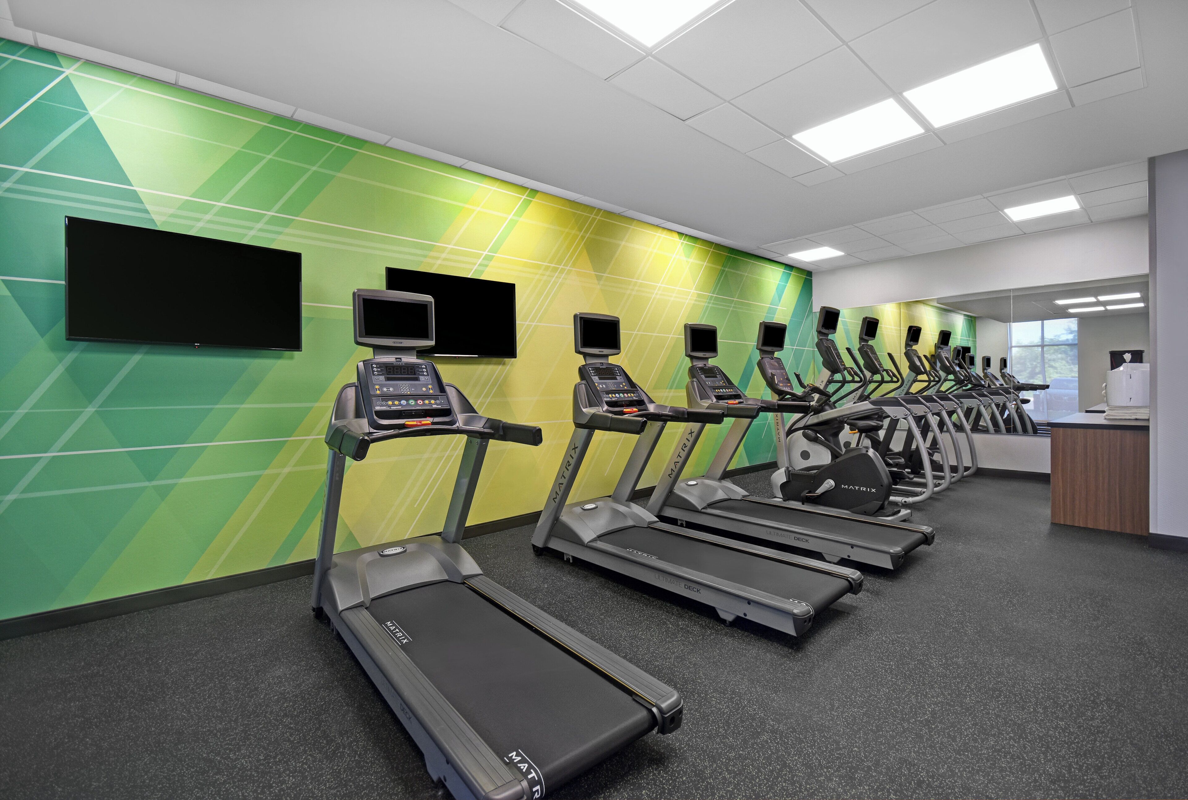 A fitness center with treadmills lined up against a green wall, a reception desk, and a window.