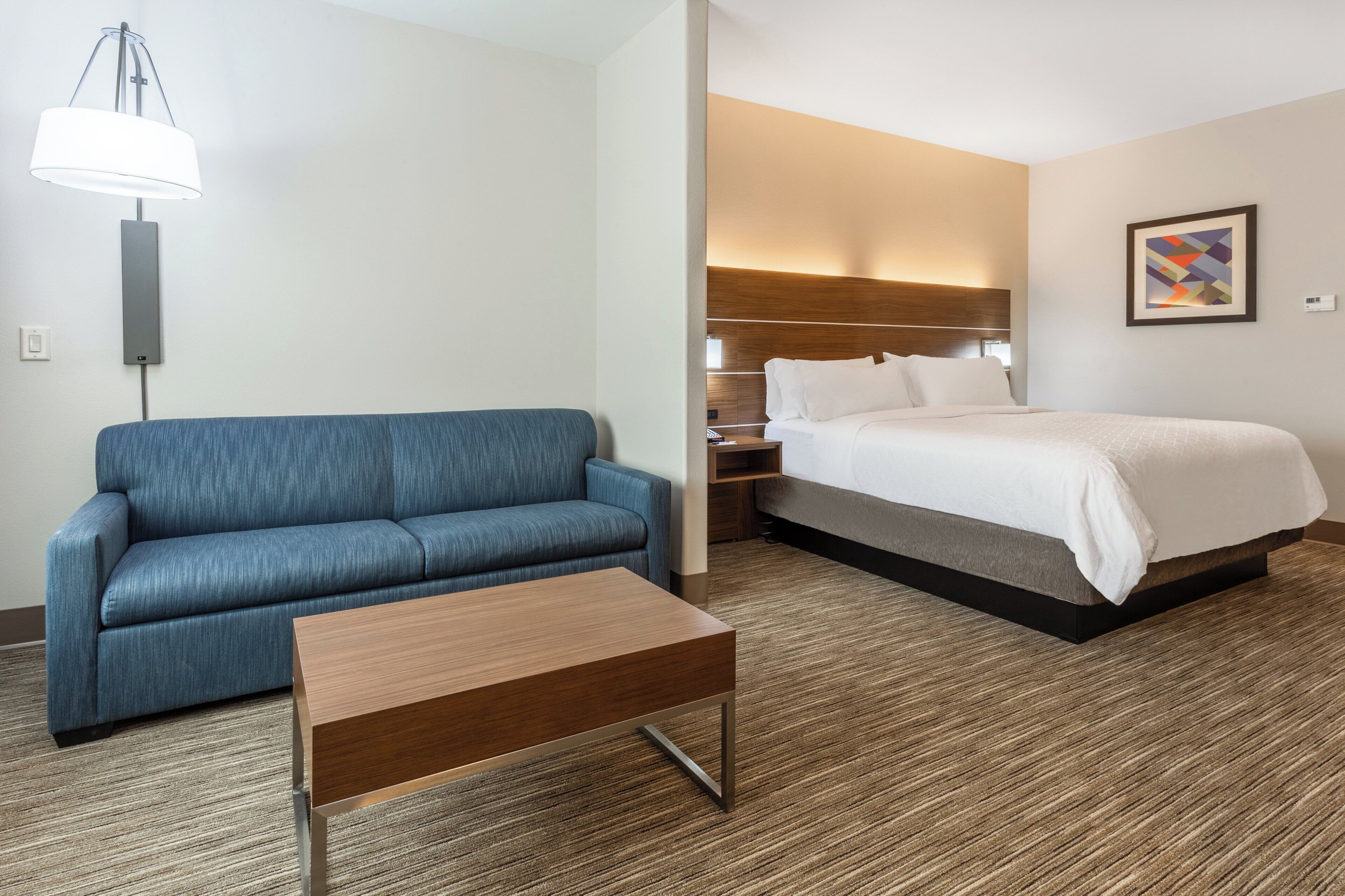 Een hotelkamer met een blauwe bank, een houten salontafel, een bed met witte beddengoed, een nachtkastje en een wandlamp.