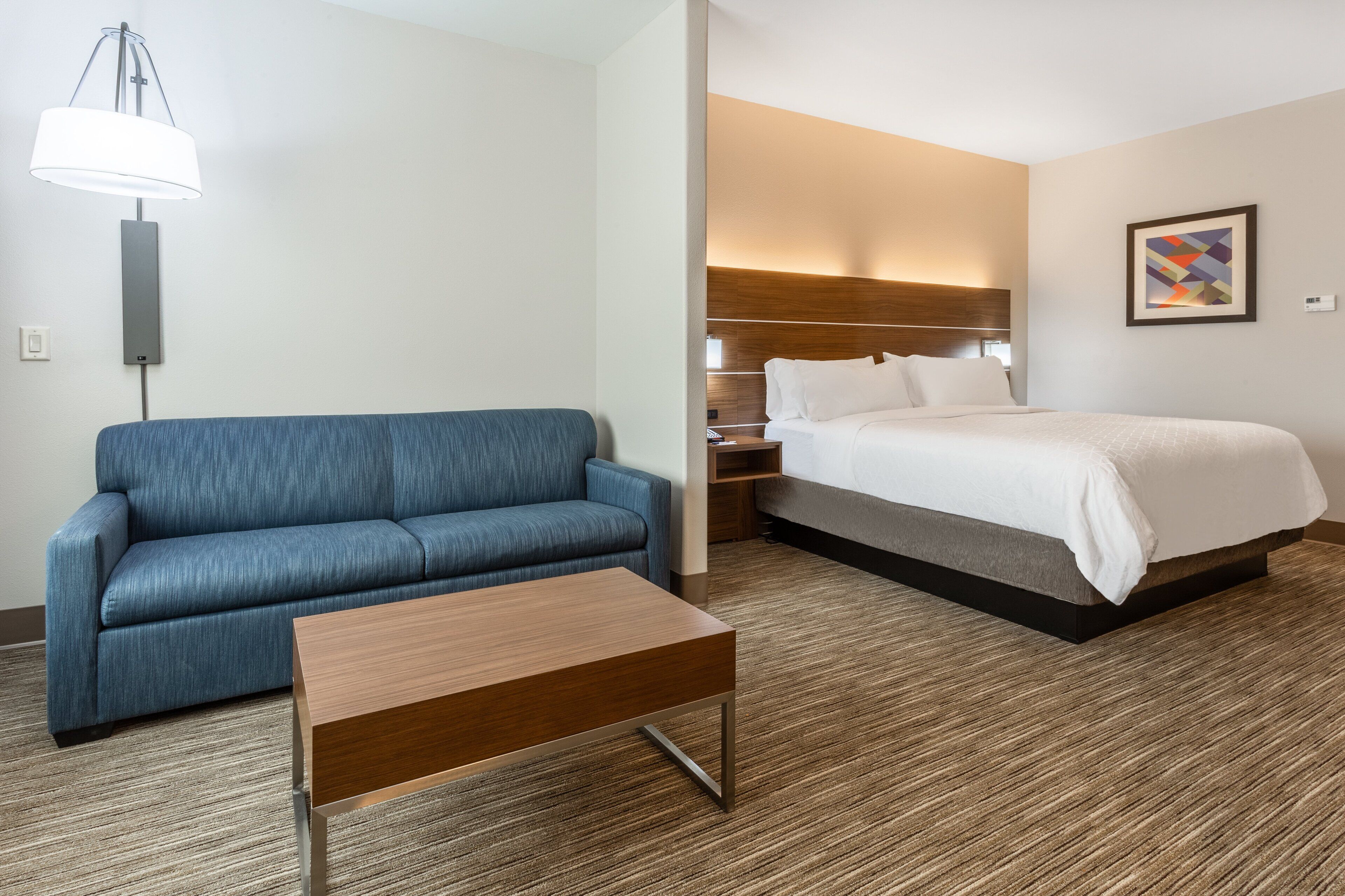 A hotel room with a blue sofa, a wooden coffee table, a bed with white bedding, a nightstand, and a wall-mounted lamp.