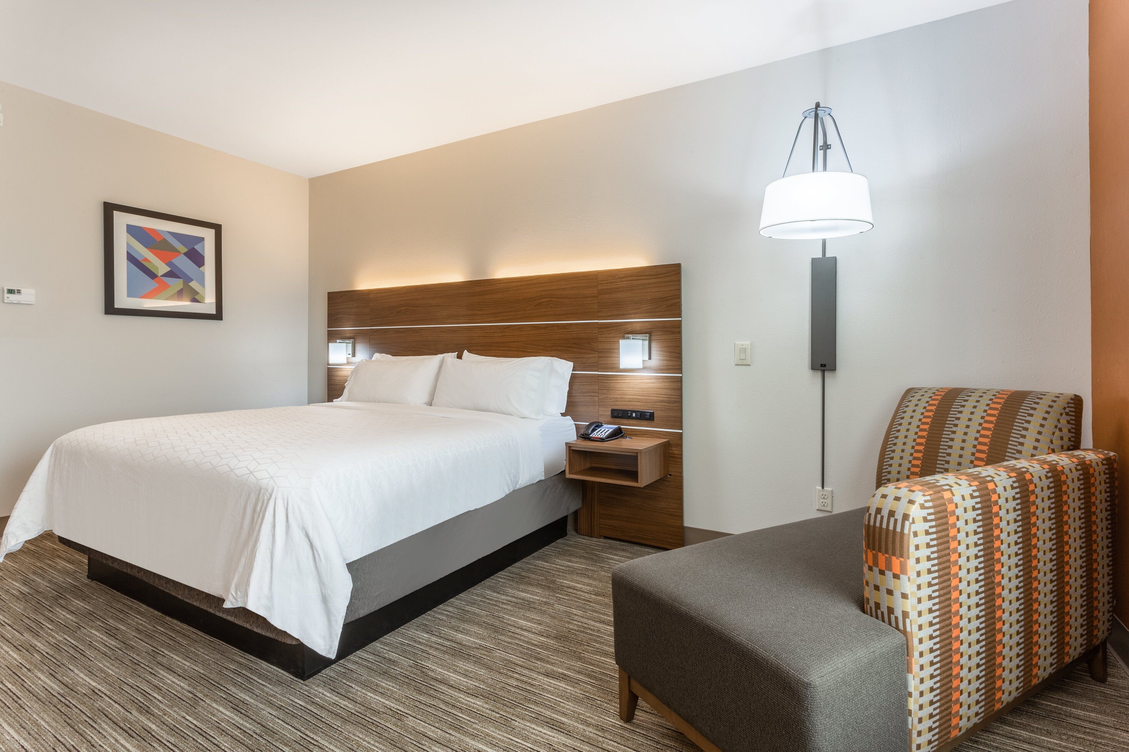 A hotel room with a bed, a nightstand, a lamp, and a patterned armchair.
