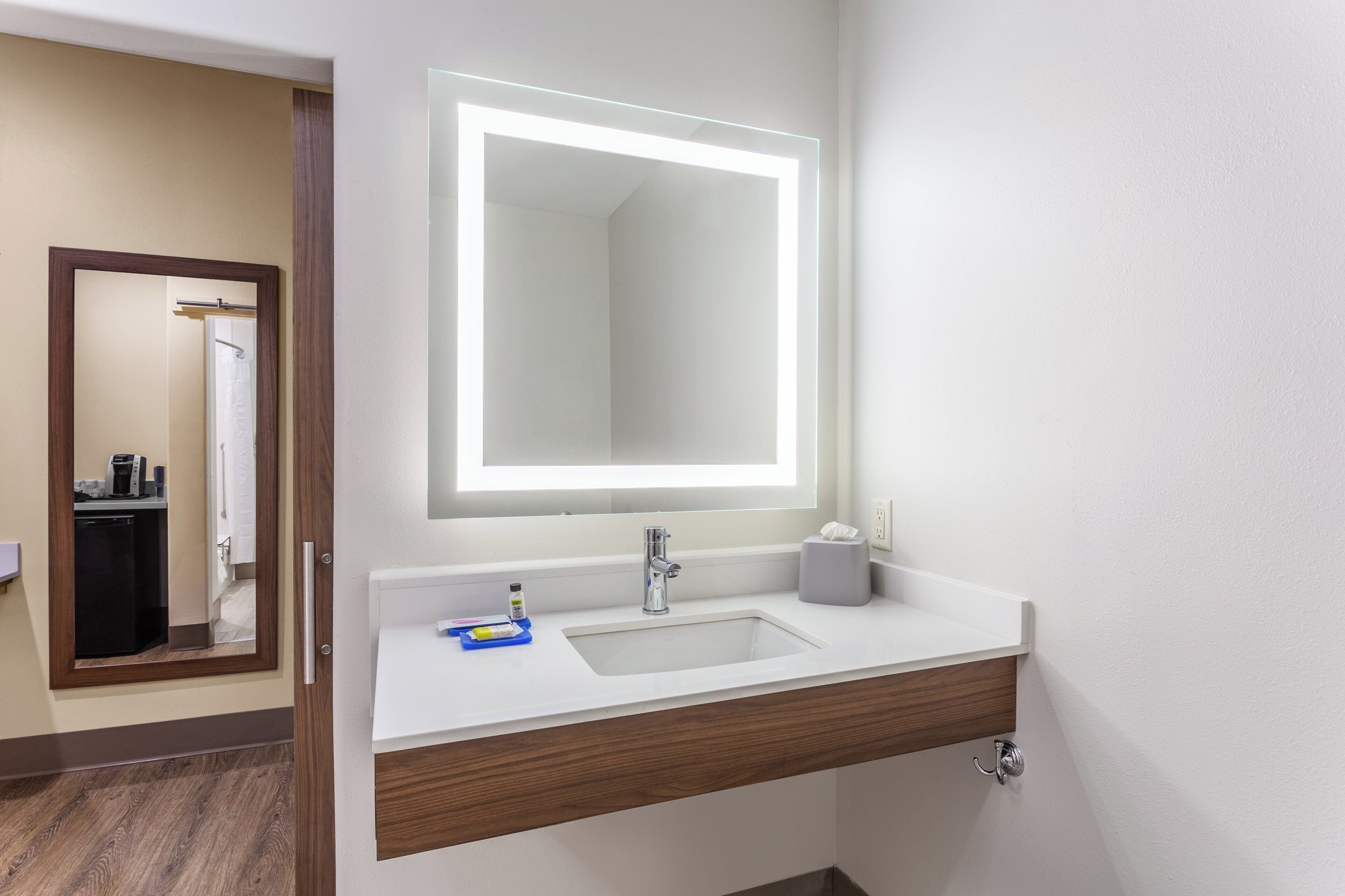 Et badeværelse på et hotel med en hvid vask, et spejl med LED-belysning, en håndklæderække og en lille håndspritflaske.