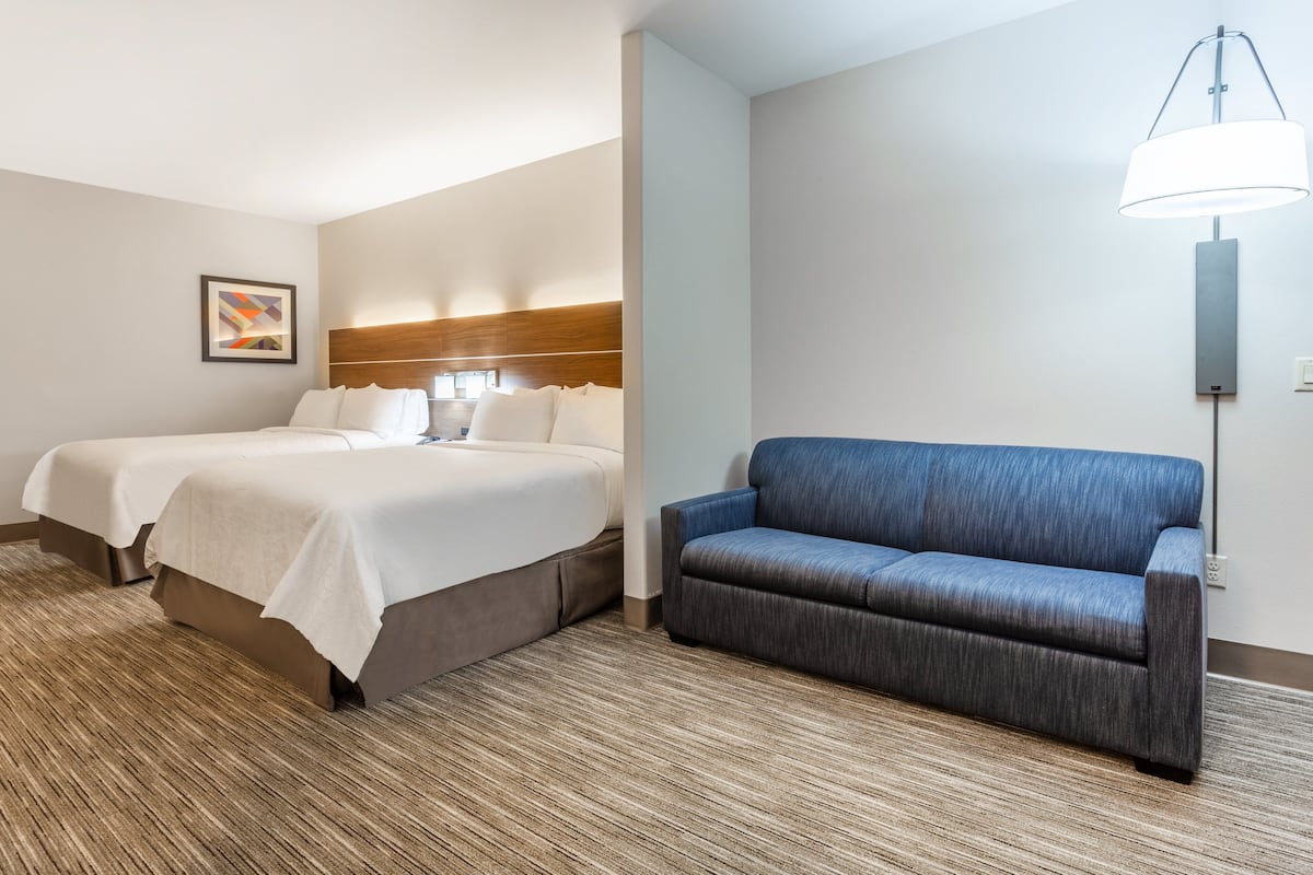 A hotel room with two beds, a blue sofa, a wooden headboard, a framed picture on the wall, and a floor lamp.