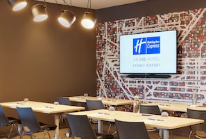 A conference room with tables, chairs, and a large screen displaying the Holiday Inn Express logo. - Holiday Inn Express Sydney Airport by IHG (Mascot)