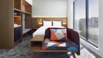 A hotel room with a bed, bedside tables, a desk, a sofa, and a large window offering a city view.
