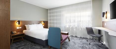 Ein Hotelzimmer mit Bett, Nachttisch, Lampe, Stuhl, Schreibtisch und einem an der Wand montierten Fernseher.