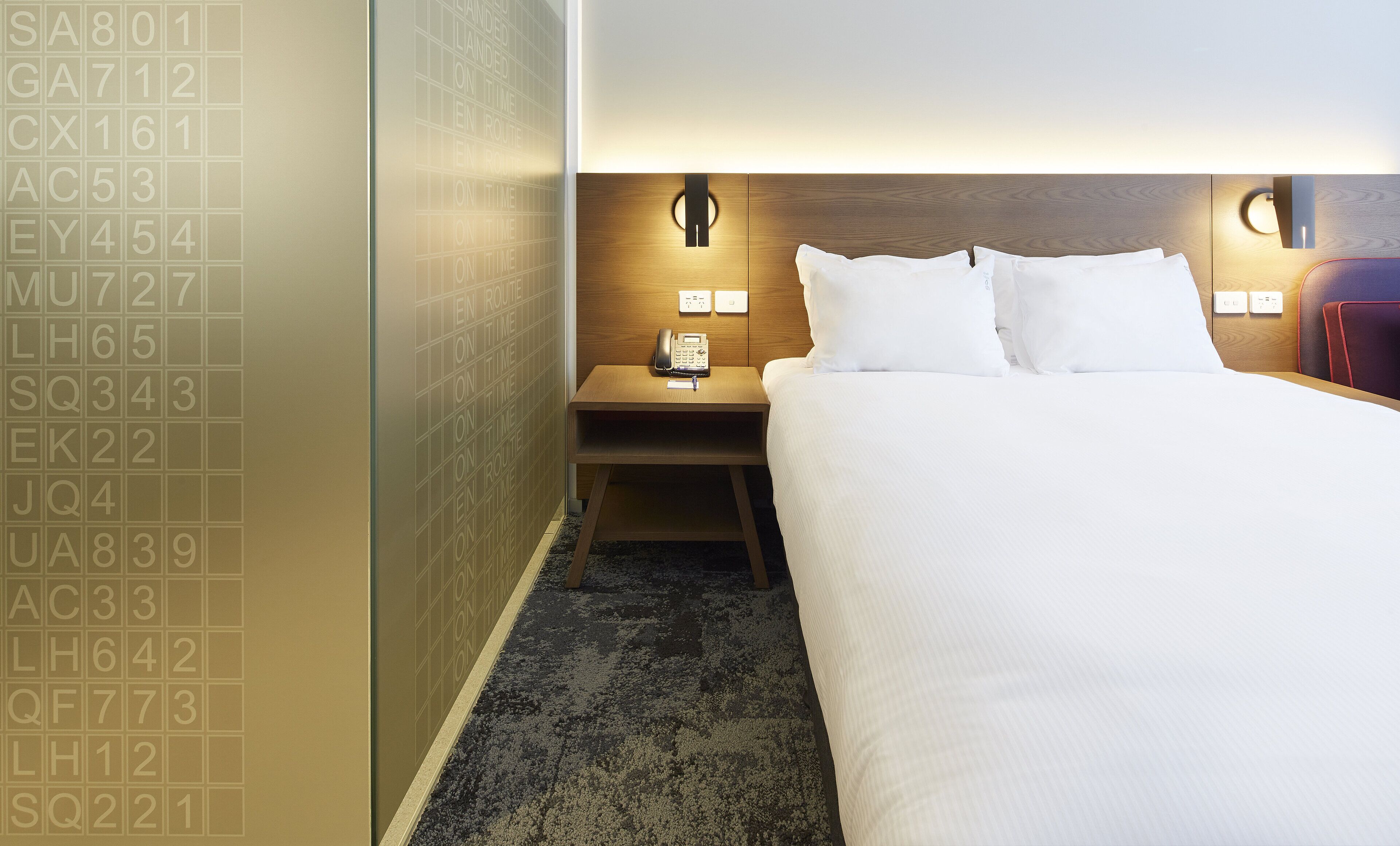 A hotel room with a neatly made bed, a bedside table with a lamp, and a wall with a grid of numbers.