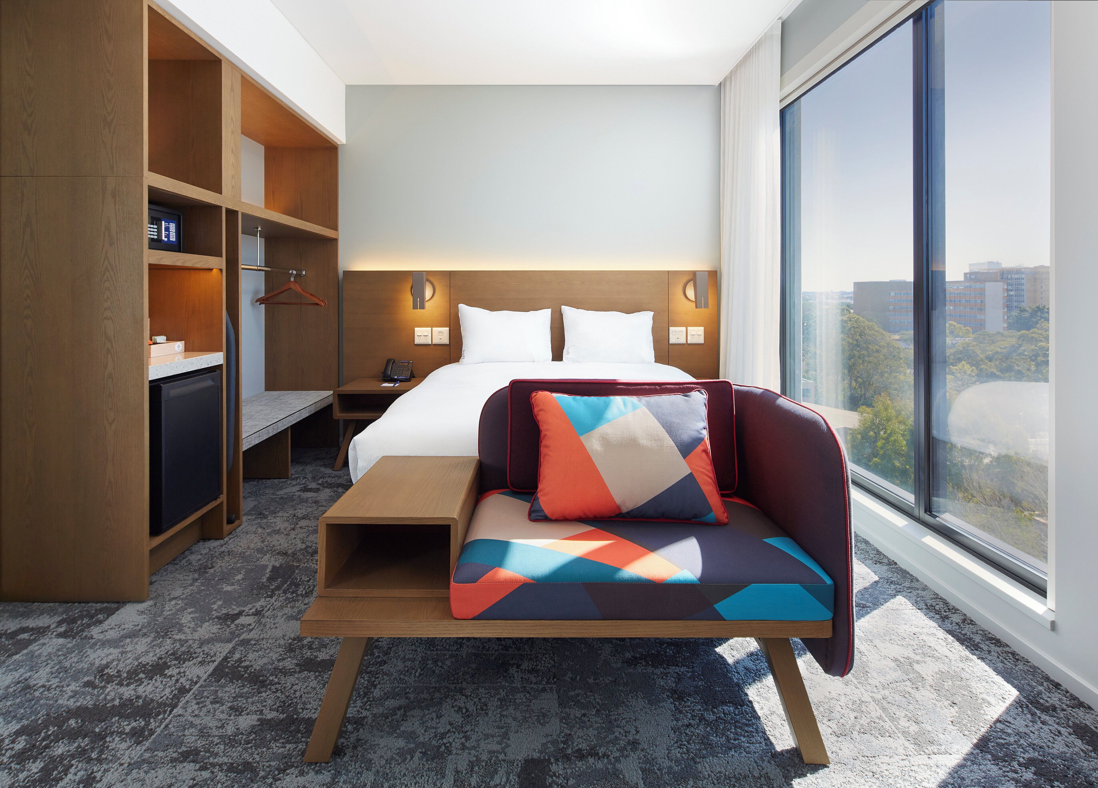 Une chambre d’hôtel équipée d’un lit, d’une table de chevet, d’un bureau, d’une armoire et d’une grande fenêtre donnant sur la ville.