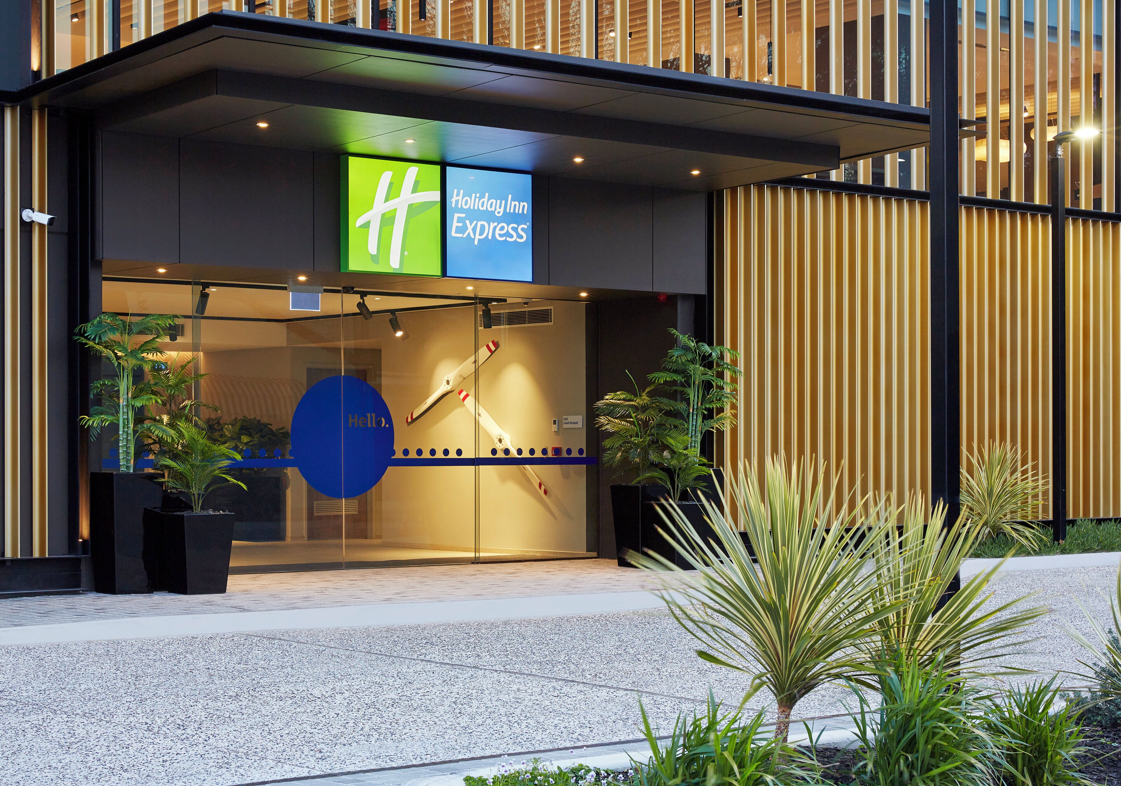 A Holiday Inn Express hotel entrance with a glass facade, security cameras, and potted plants.