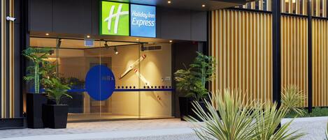 Entrada de un hotel Holiday Inn Express con fachada de vidrio, cámaras de seguridad y plantas en macetas.