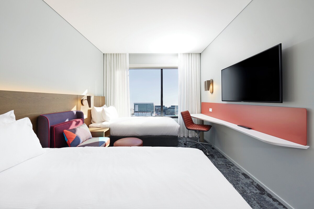 Een hotelkamer met een groot bed, een bureau, een stoel, een flatscreen-televisie en een raam met uitzicht op de stad.