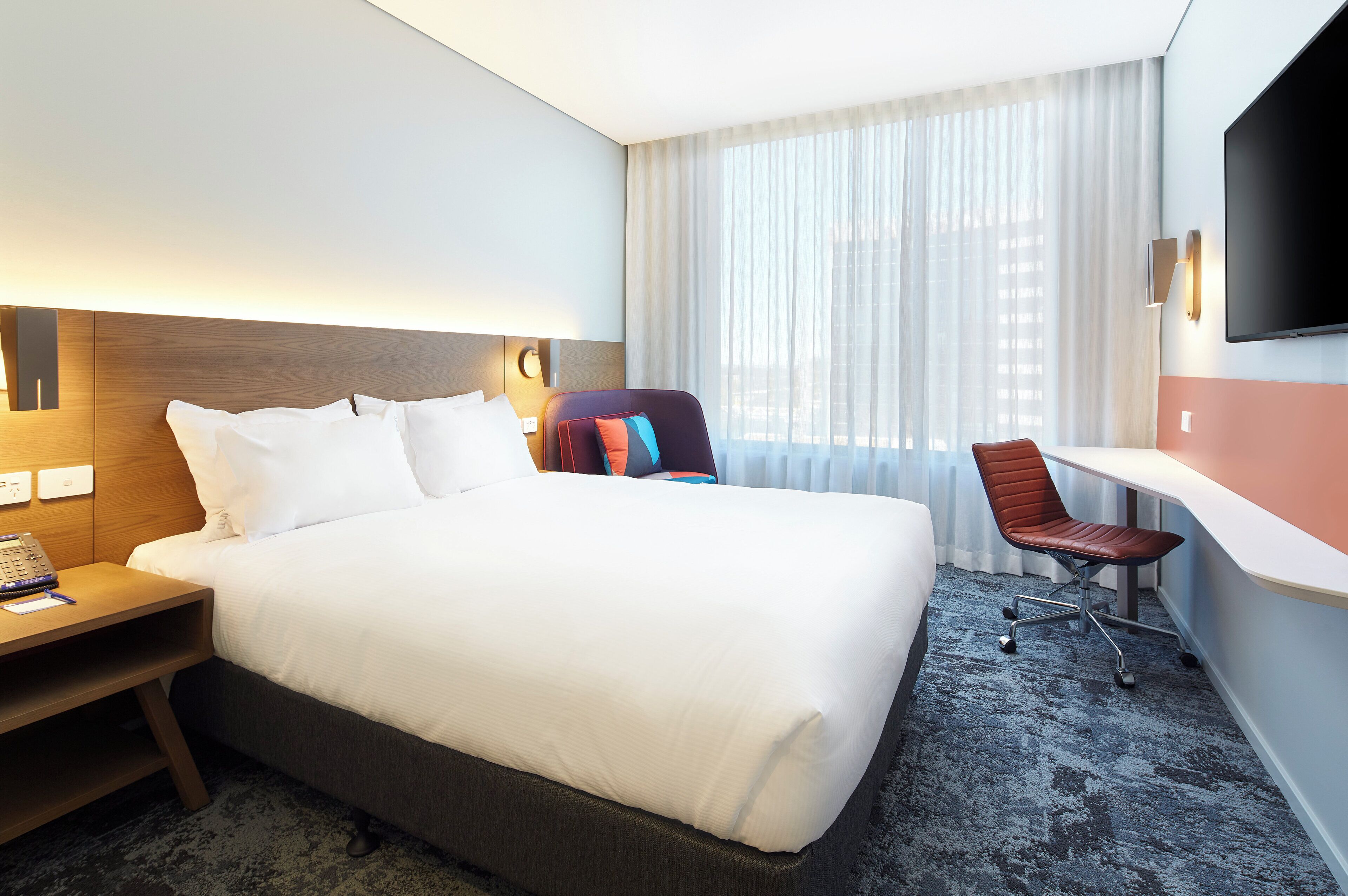 A hotel room with a large bed, a desk with a chair, a flat-screen TV, and a window with curtains.