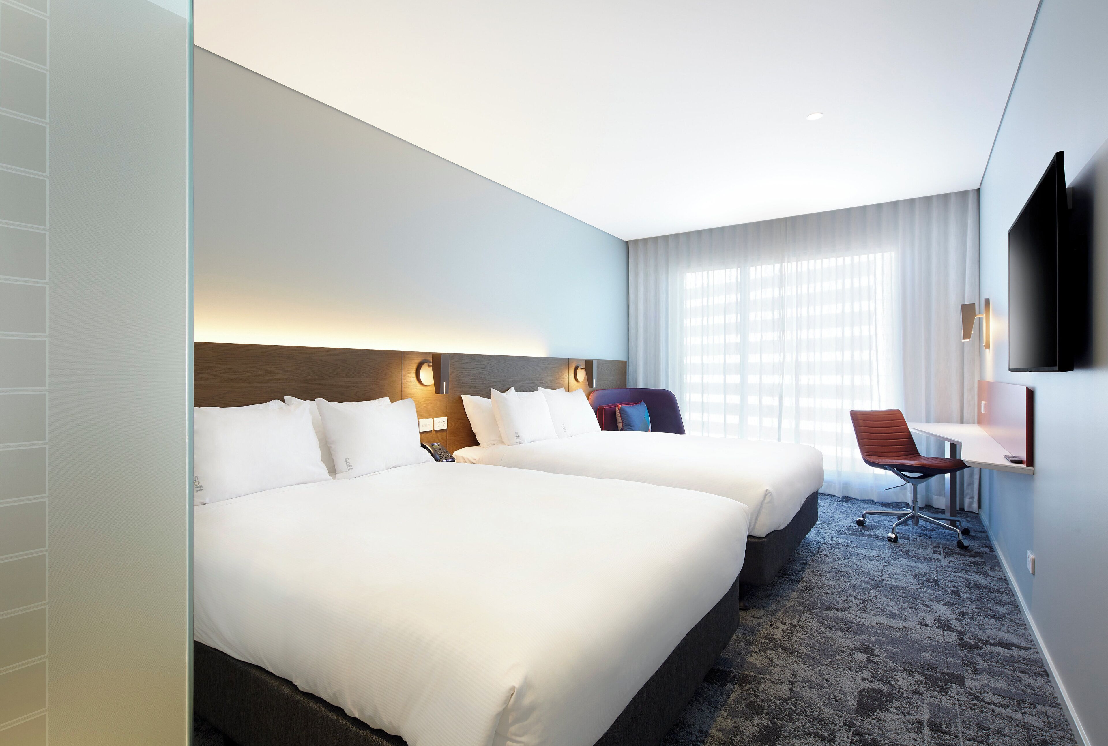 A hotel room with two beds, a desk, a chair, a flat-screen TV, and a large window with curtains.